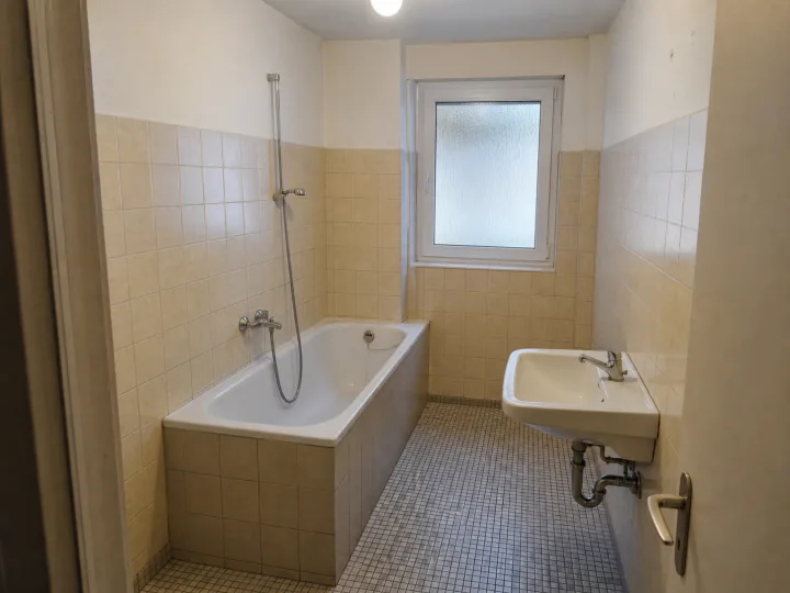 Schmales Bad mit Badewanne, beigefarbenen Wandfliesen, kleinteiligem Mosaikboden und Waschbecken
