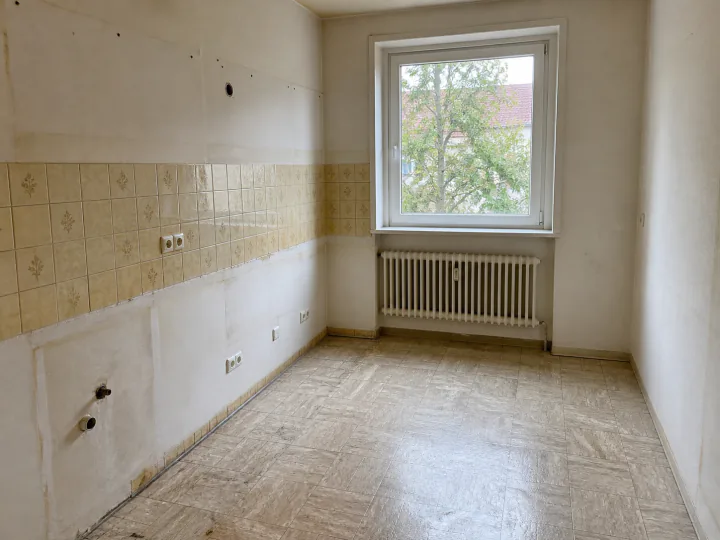 Leerer Küchenraum mit gemustertem Fliesenspiegel, offenen Wasseranschlüssen, Steckdosen, Heizkörper und Fenster