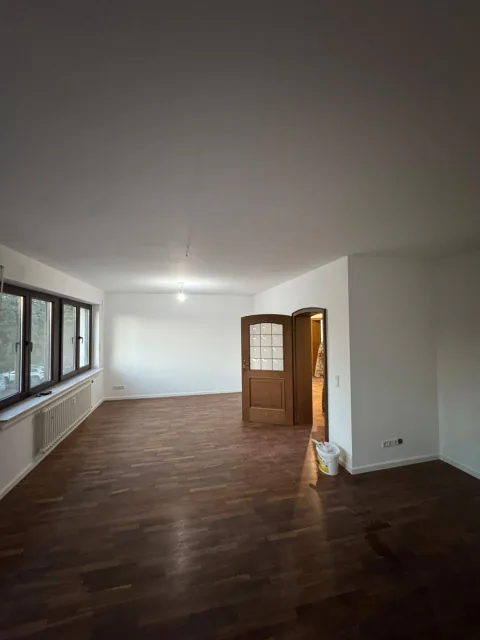 Großer renovierter Wohnraum mit weißer Decke, dunklem Boden in Holzoptik, Fensterfront und brauner Tür