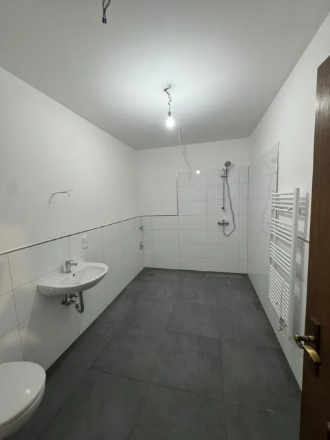 Helles Badezimmer mit weißen Wandfliesen, grauem Boden, Waschbecken, Wand-WC und Duschanschlüssen